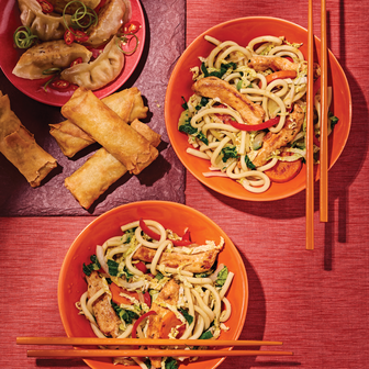 Two bowls of stir-fried noodles with vegetables and chicken, served with chopsticks, alongside spring rolls and dumplings on a red background.